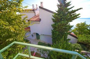 A white building with a red-tiled roof, surrounded by greenery and overlooking a body of water.