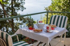 A table set with a teapot, cups, and pastries on a balcony with chairs, overlooking a body of water.