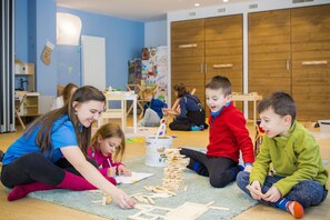 A group of children engaged in a building activity with wooden blocks on a carpeted floor in a room with blue walls and wooden cabinets.