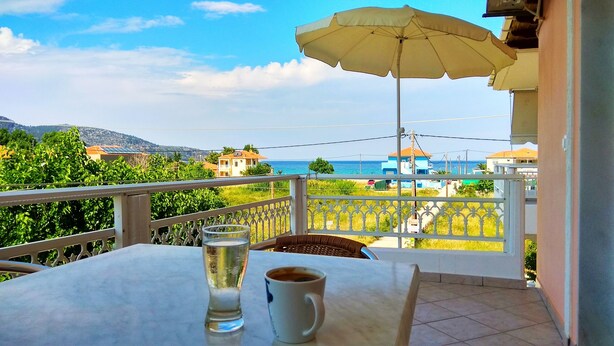A balcony with a table, a glass of water, and a cup of coffee. A yellow umbrella is set up for shade. The view includes a body of water and buildings in the distance. - Porto Thassos (Thasos)