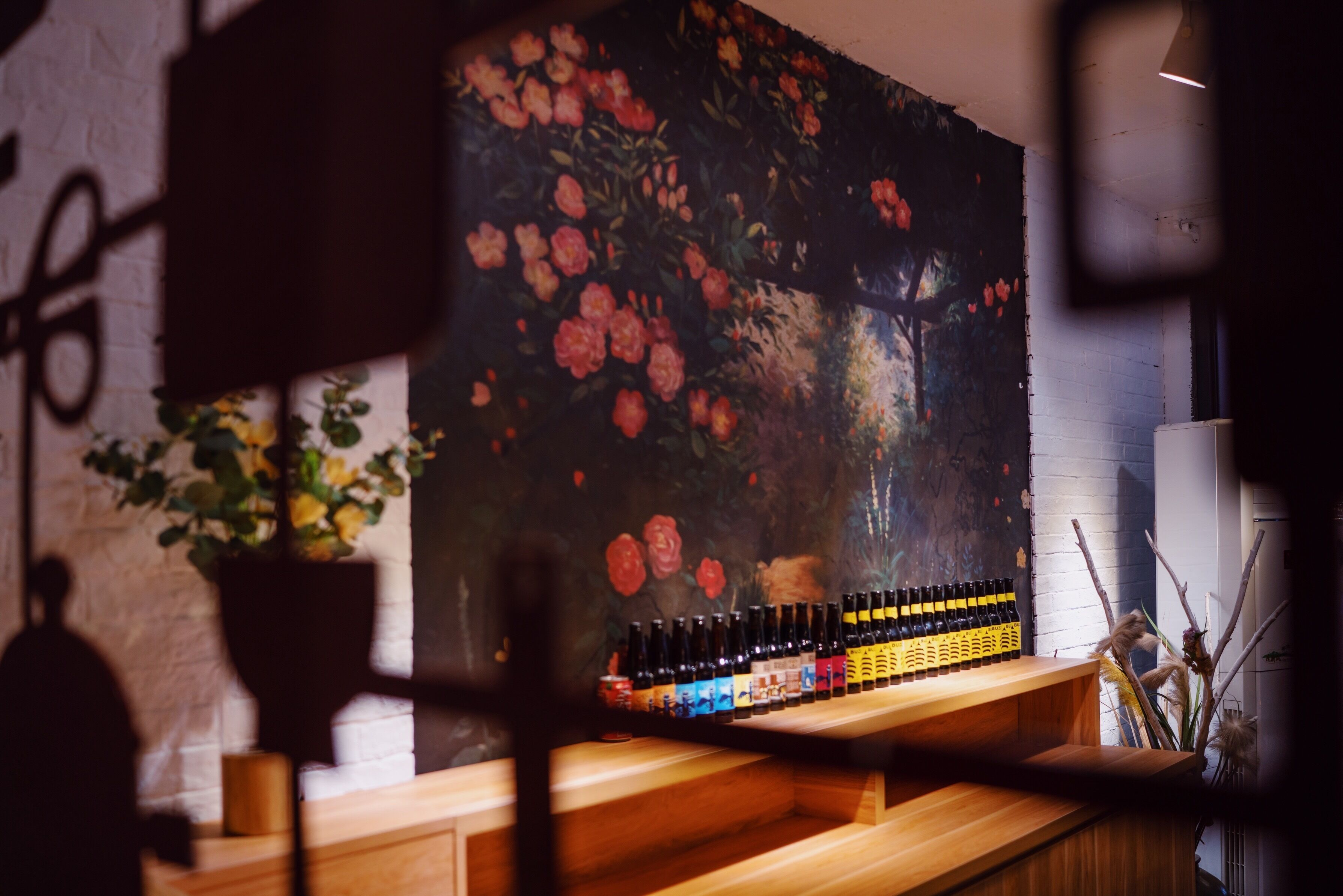 Uma área de bar com balcão de madeira, um grande mural floral na parede e garrafas de bebidas.