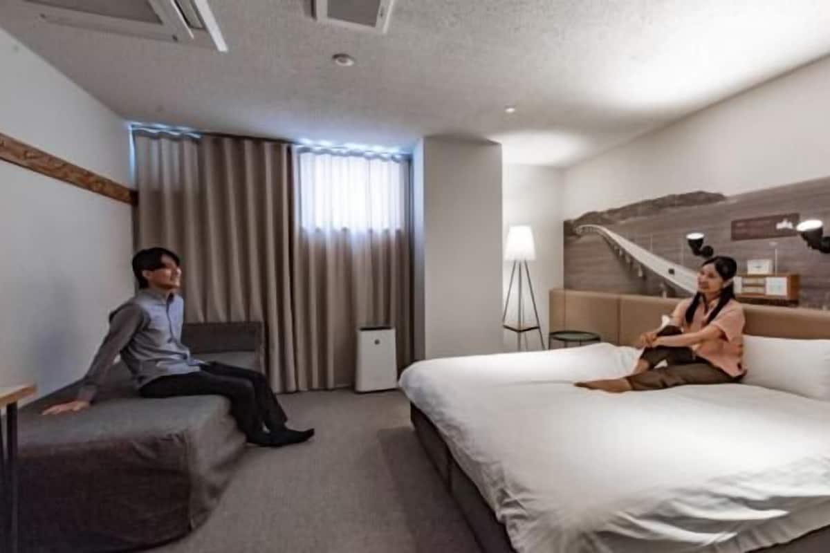 A hotel room with a bed, a desk, a chair, a standing lamp, and a window with curtains.