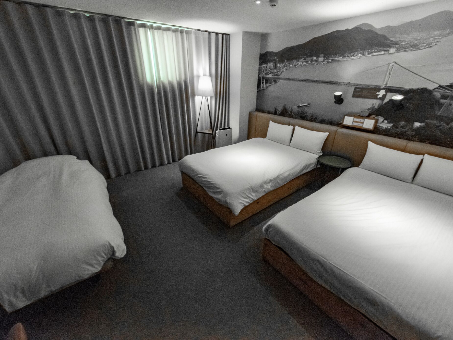 A hotel room with two single beds, a large window with curtains, a nightstand with a lamp, and a mural of a bridge and mountains on the wall.