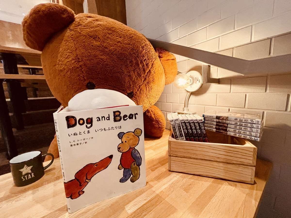 Ein gemütliches Zimmer mit einem großen Teddybären, einem Buch mit dem Titel 'Hund und Bär', einer Holzkiste und einer Star-Tasse auf dem Tisch.