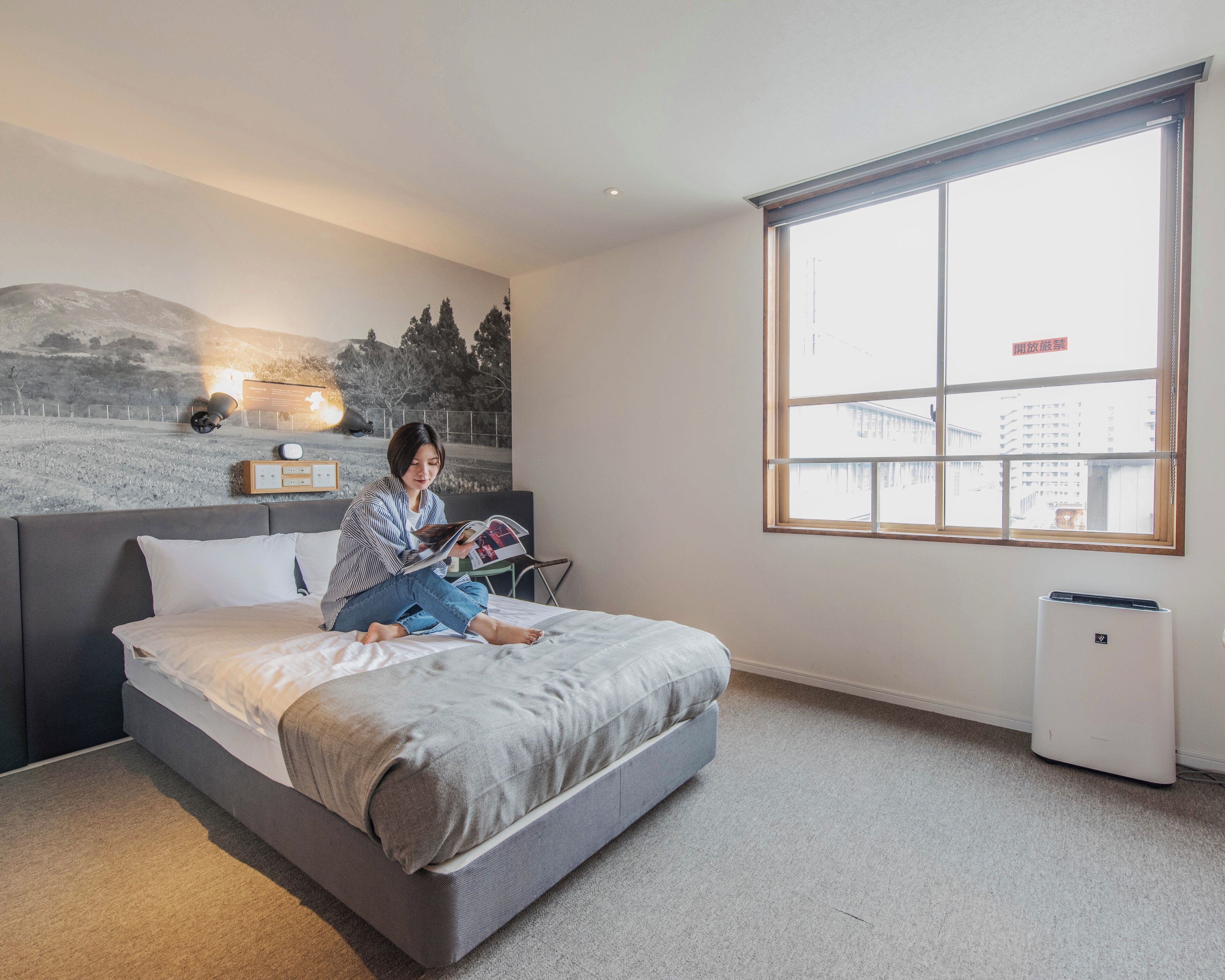 A modern bedroom with a large bed, a wall-mounted reading light, a window with a view of buildings, and a standing air conditioner.