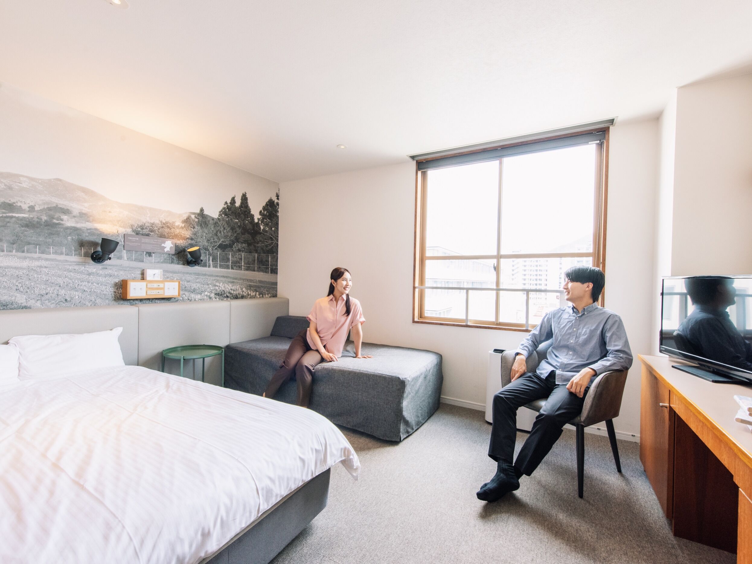 Habitación de hotel con cama, sofá, escritorio con televisor y ventana grande.