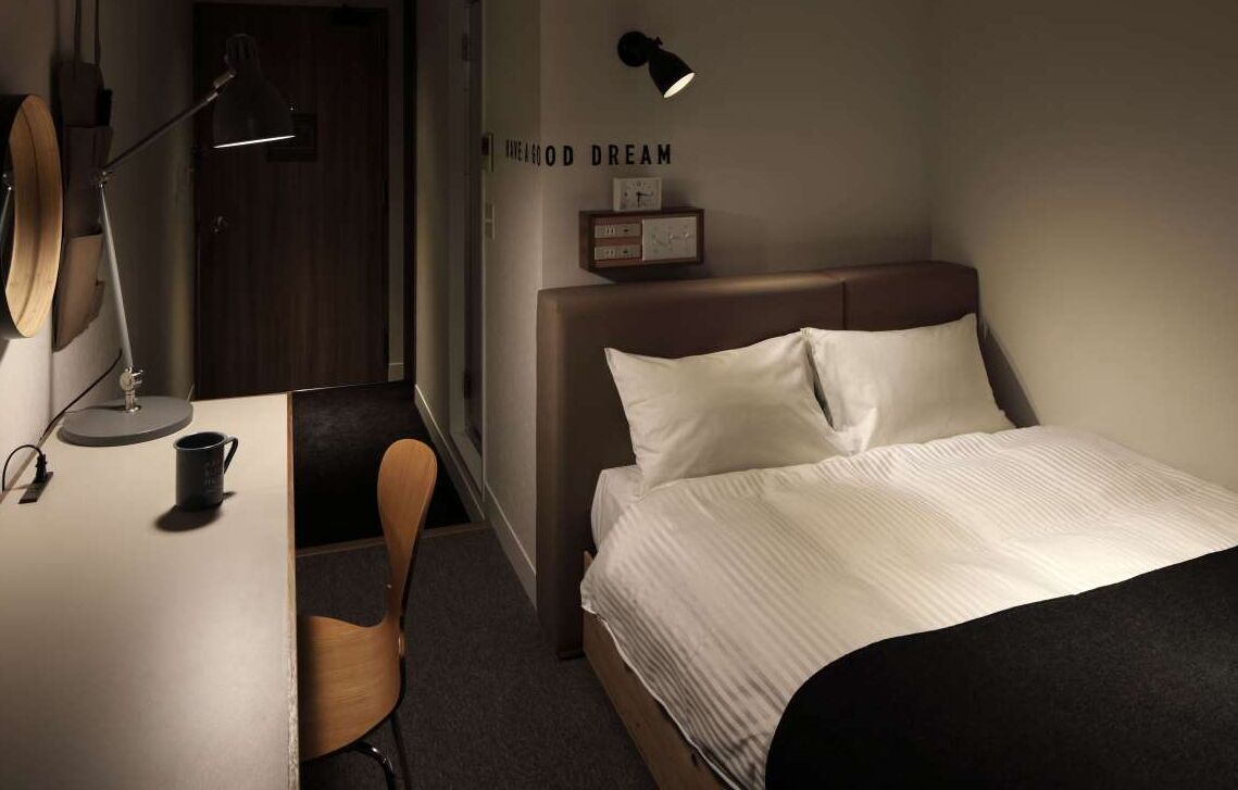 A neatly arranged hotel room with a bed, a desk, a chair, and a desk lamp.
