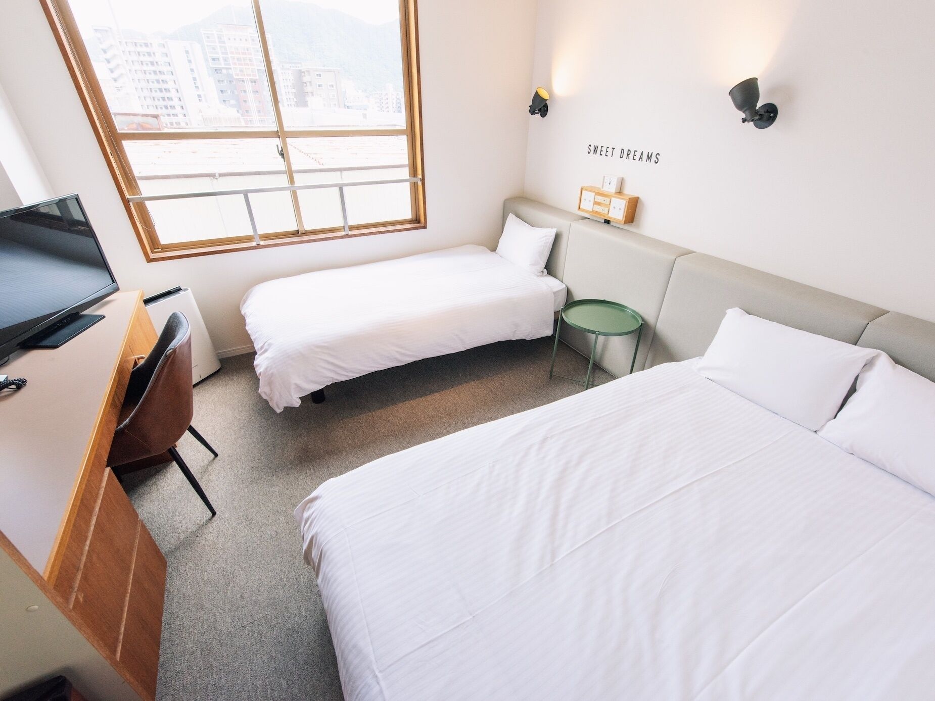 Habitación de hotel con dos camas individuales, un escritorio con computadora, una mesita pequeña y un estante en la pared.
