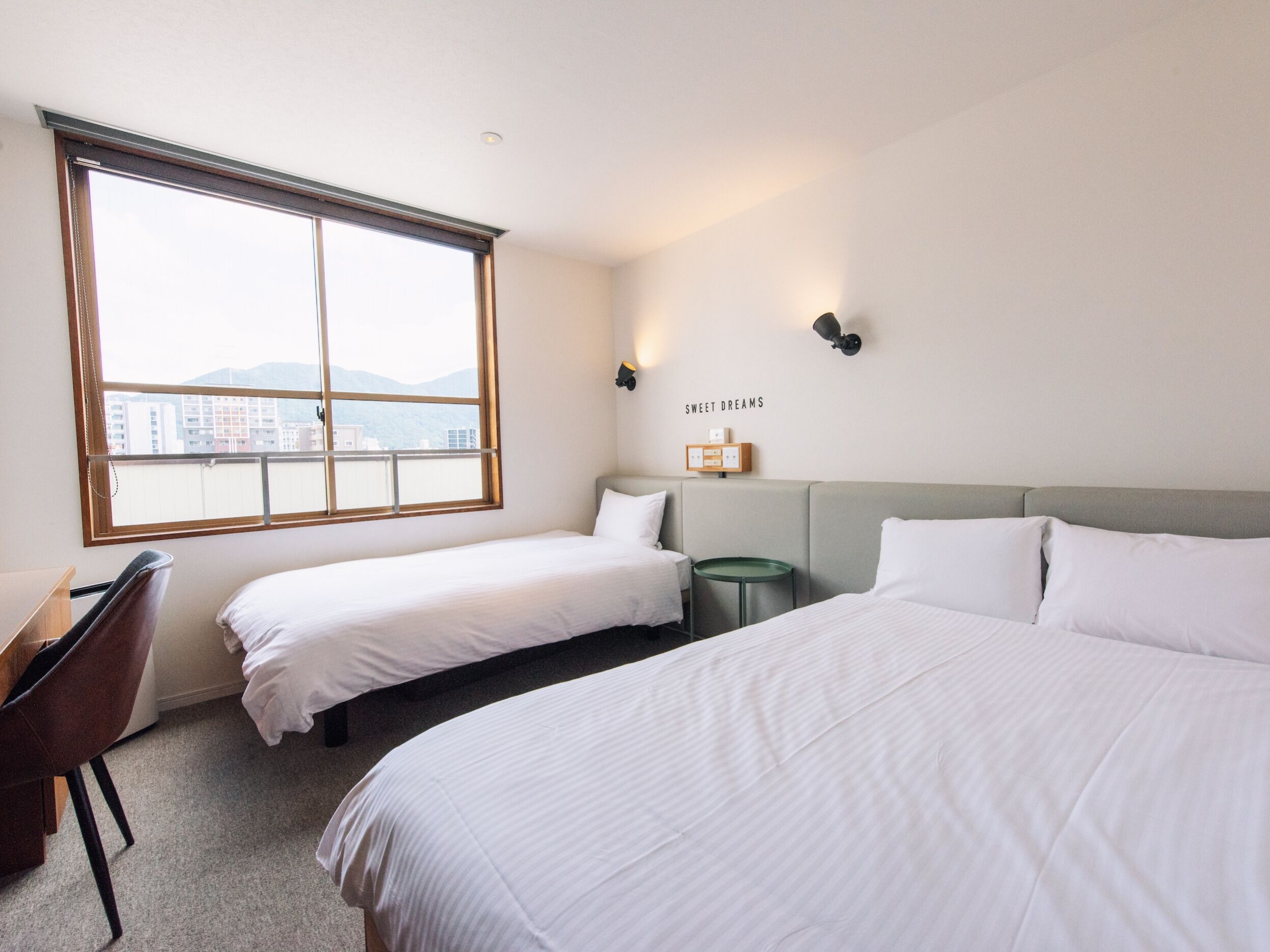 Habitación de hotel con dos camas individuales, un escritorio y una silla. Amplia ventana con vistas a edificios y montañas.