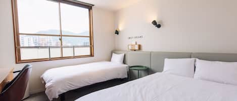 A hotel room with two single beds, a desk, and a chair. There is a large window providing a view of buildings and mountains.