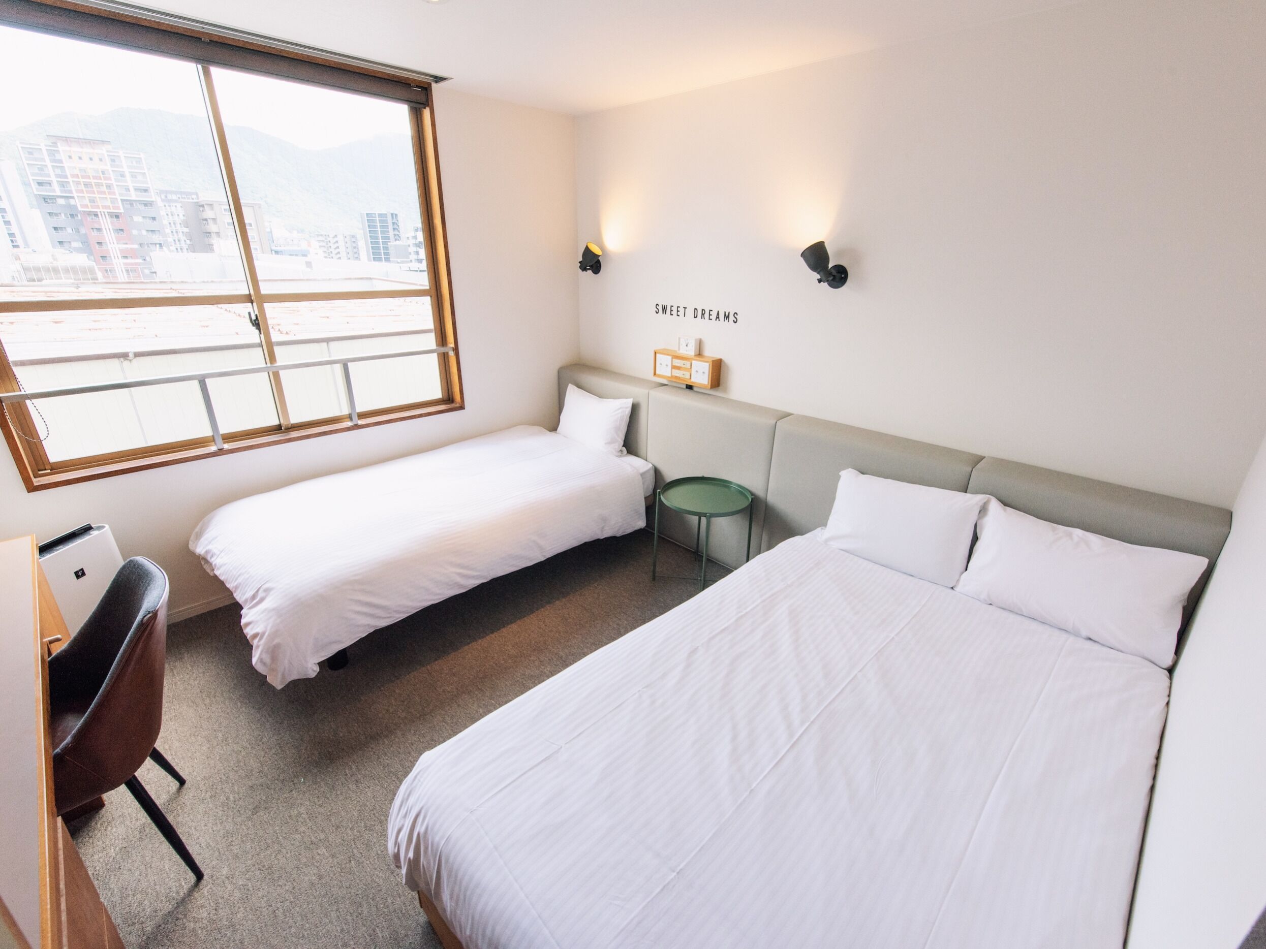 A hotel room with two beds, a desk, a chair, a small table, and a window with a view of buildings and mountains.