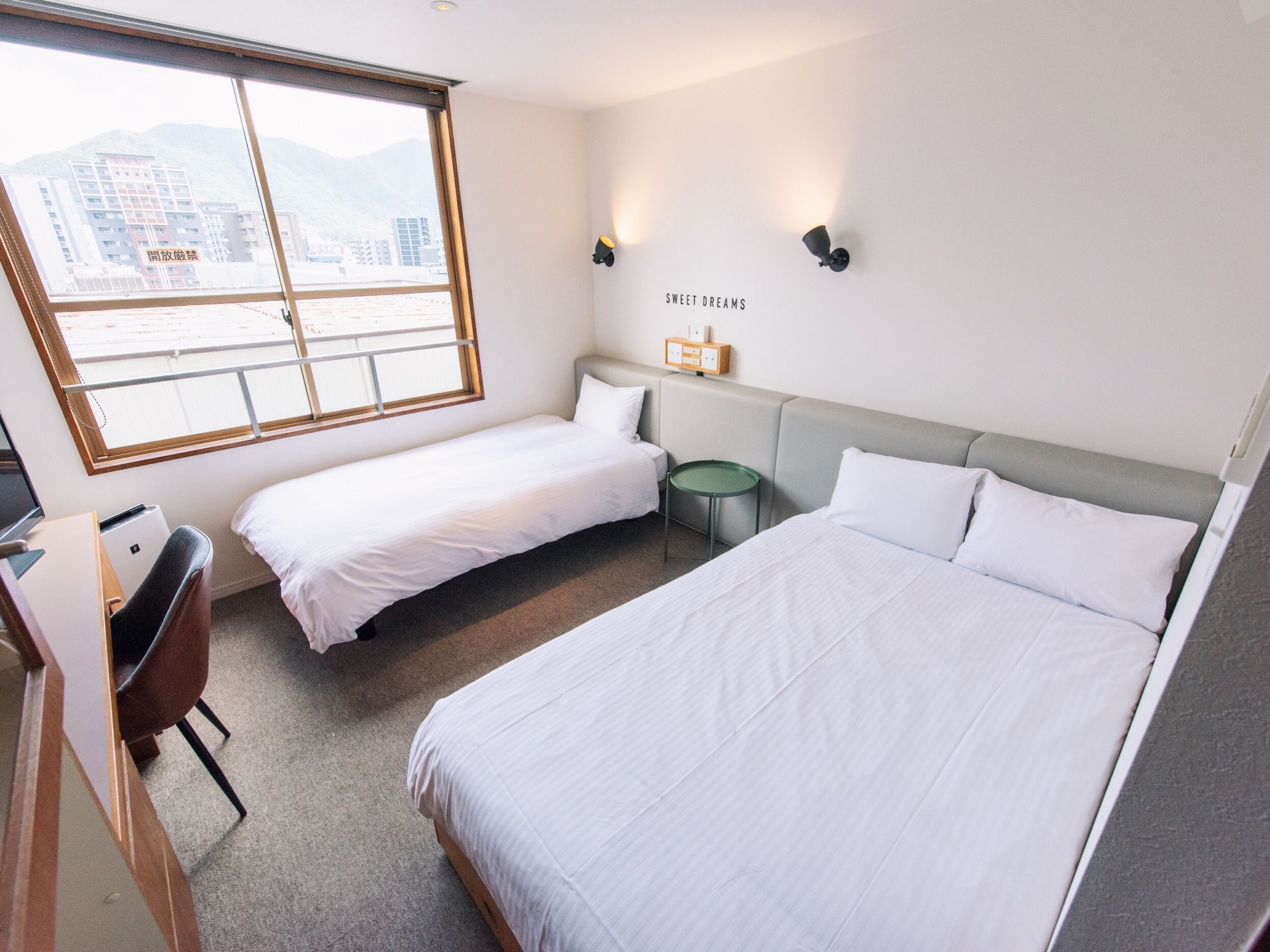 A hotel room with two single beds, a desk, a chair, a small round table, and a wall-mounted lamp.