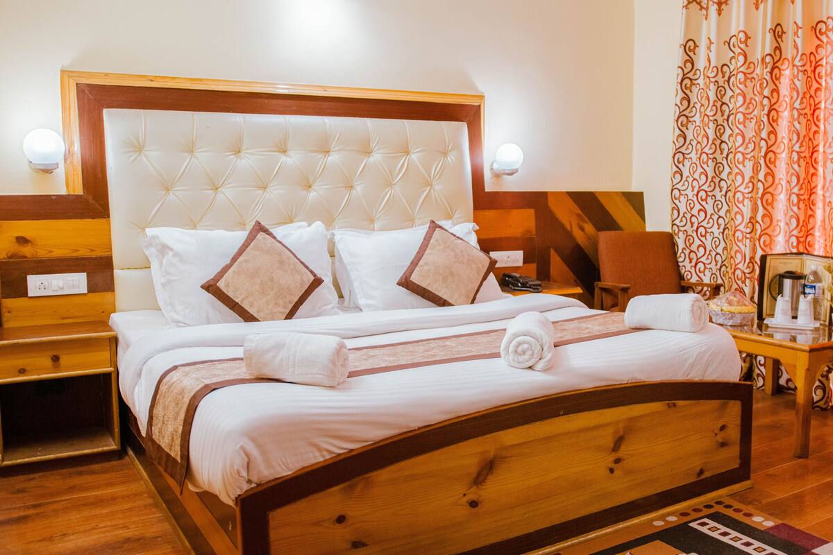 A neatly made bed with a wooden headboard, white bedding, and two brown pillows. There is a wooden nightstand with a lamp, a chair, and a small table with a coffee maker and a plant.