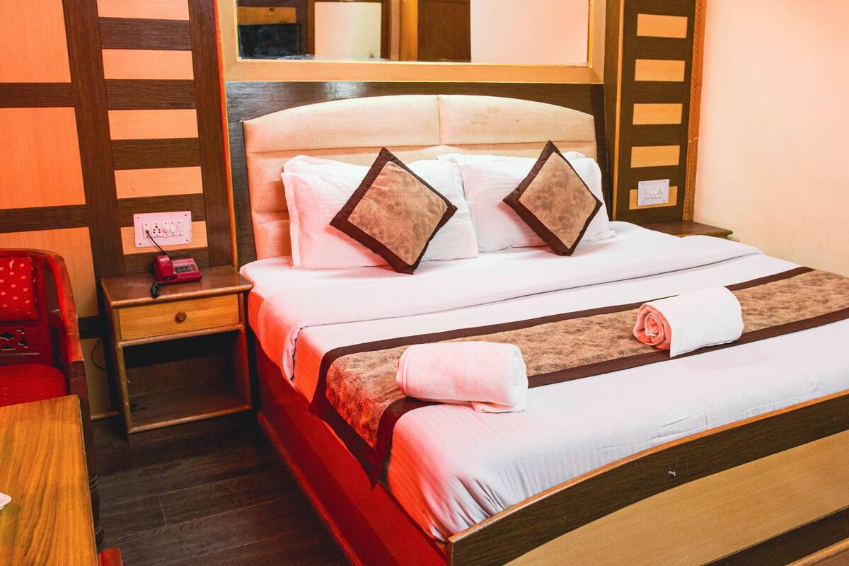 A neatly made bed with white linens and two decorative pillows, a wooden nightstand with a red chair, and a wall-mounted mirror.