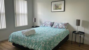 Une chambre à coucher comprenant un lit, deux tables de chevet, une lampe, une fenêtre avec des stores et un tableau encadré accroché au mur.