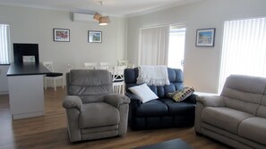 Un salon comprenant un canapé, des fauteuils, une table basse et un coin repas avec des chaises et une table. Les murs sont ornés de tableaux encadrés et un téléviseur est posé sur un meuble.