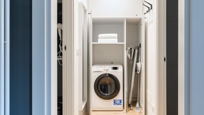 Ein Waschraum mit einer Frontlader-Waschmaschine, einem Regal mit gefalteten Handtüchern und einem Wäscheständer.