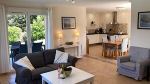 Un salon comprenant un canapé, une table basse et un coin repas avec des chaises et une table. Au fond, on aperçoit une cuisine avec un four et des placards.