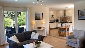 Ein Wohnzimmer mit einer Couch, einem Couchtisch und einem Essbereich mit Stühlen und einem Tisch. Im Hintergrund ist eine Küche mit einem Herd und Schränken zu sehen.