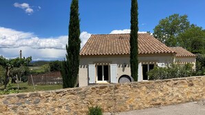 Une maison aux murs de pierre et au toit de tuiles, deux cyprès majestueux et un ciel dégagé.