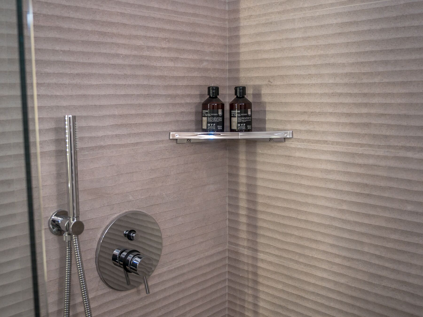 A shower area with a handheld showerhead, a wall-mounted faucet, and a shelf with two bottles.