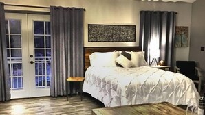 A hotel room with a bed, a desk with a glass, a chair, and a door with curtains. - Papillon Cellars (Mariposa)