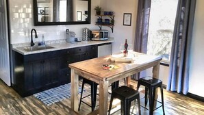 A modern kitchen with dark cabinetry, a white countertop, a wooden dining table with stools, a large mirror, and a window with curtains. - Papillon Cellars (Mariposa)