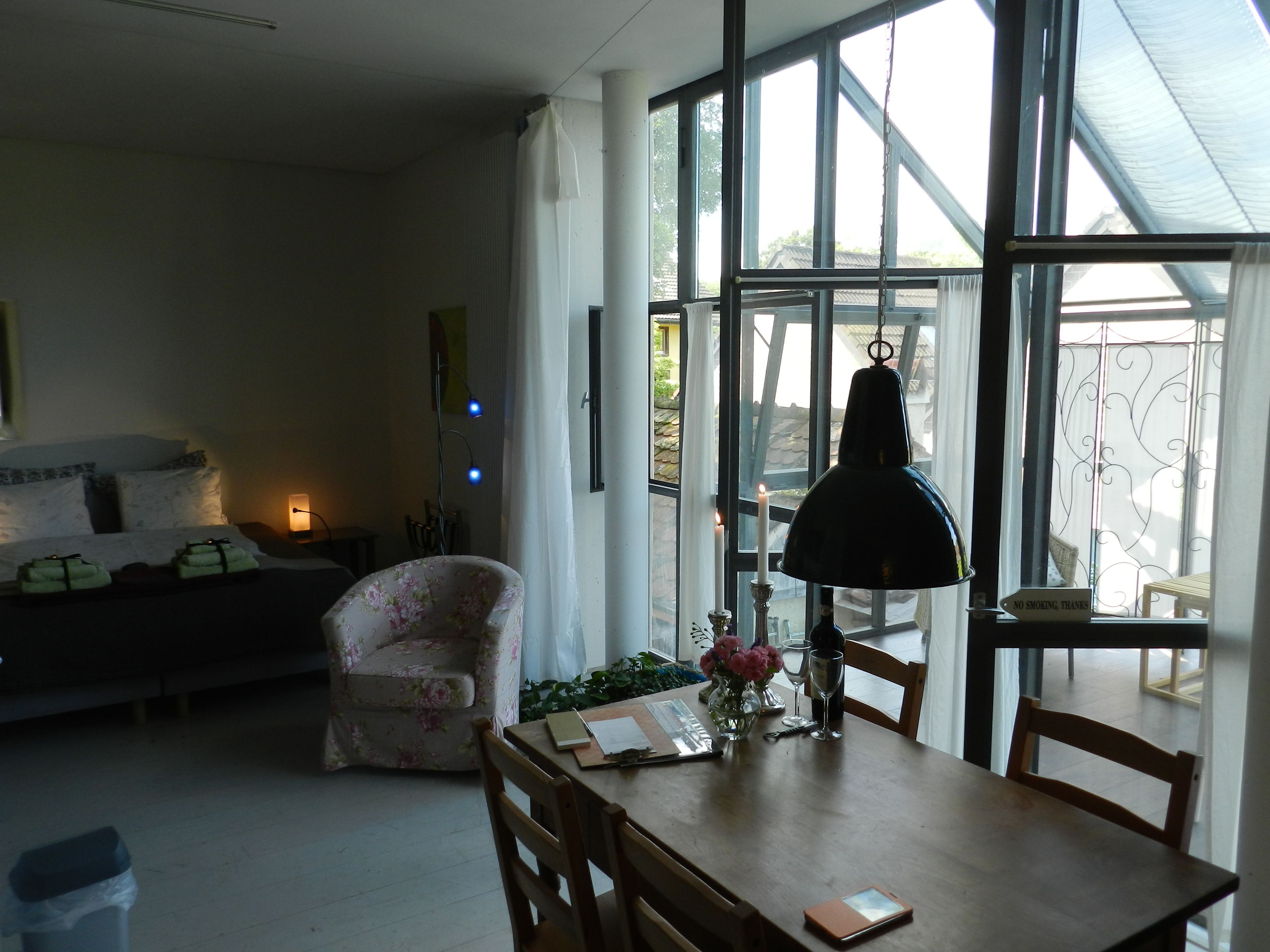 Una habitación con una cama, un sillón floral, una mesa de comedor con sillas, una lámpara y vistas amplias a través de ventanas grandes.