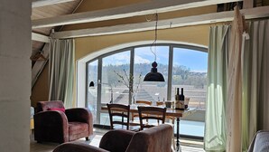 Un vaste espace de vie doté d’un plafond haut, de poutres en bois, d’une grande fenêtre avec des rideaux et d’une table à manger avec des chaises.