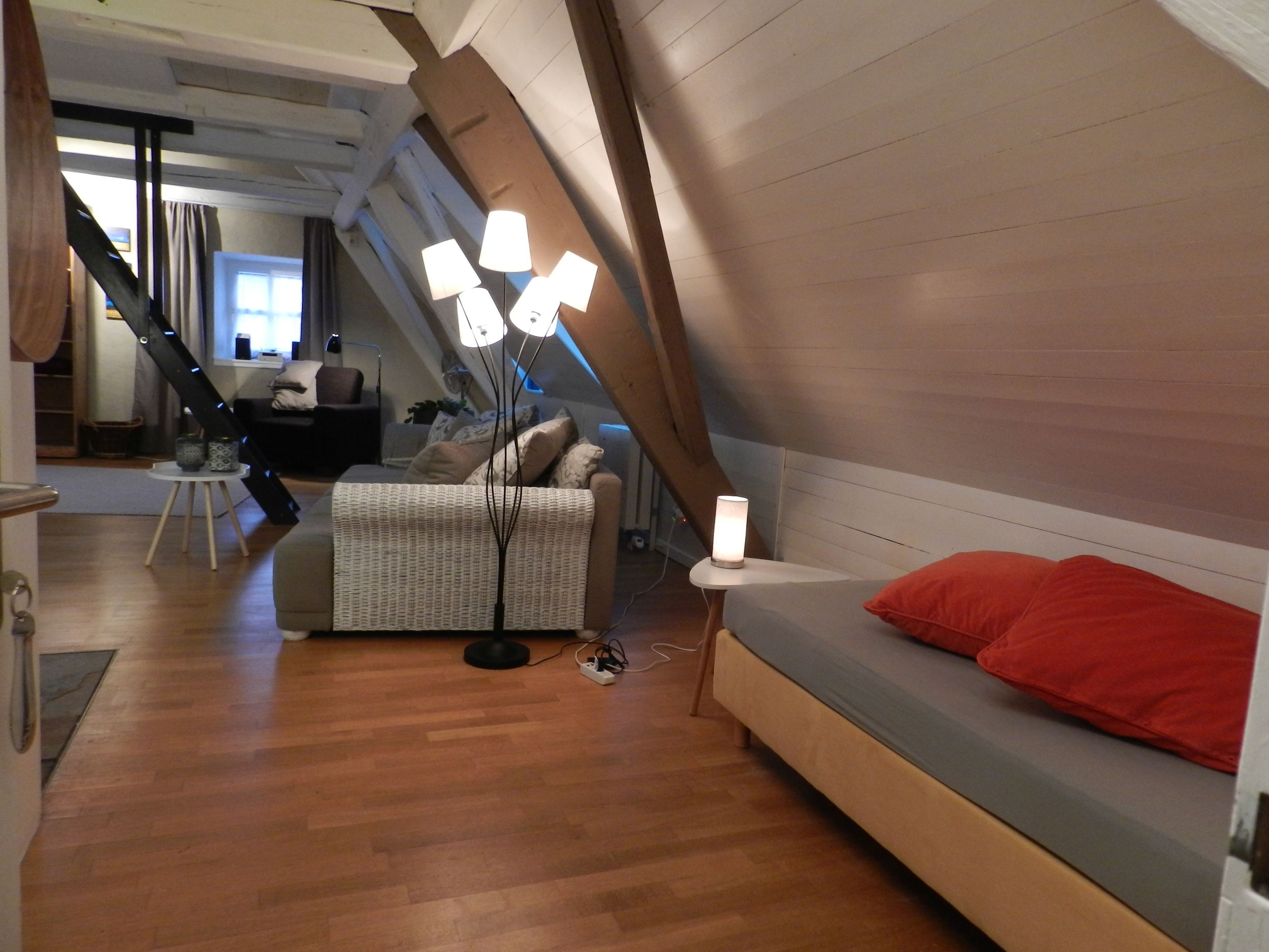 A modern loft bedroom with a sloped ceiling, a wooden floor, a sofa bed with red cushions, and a standing floor lamp.