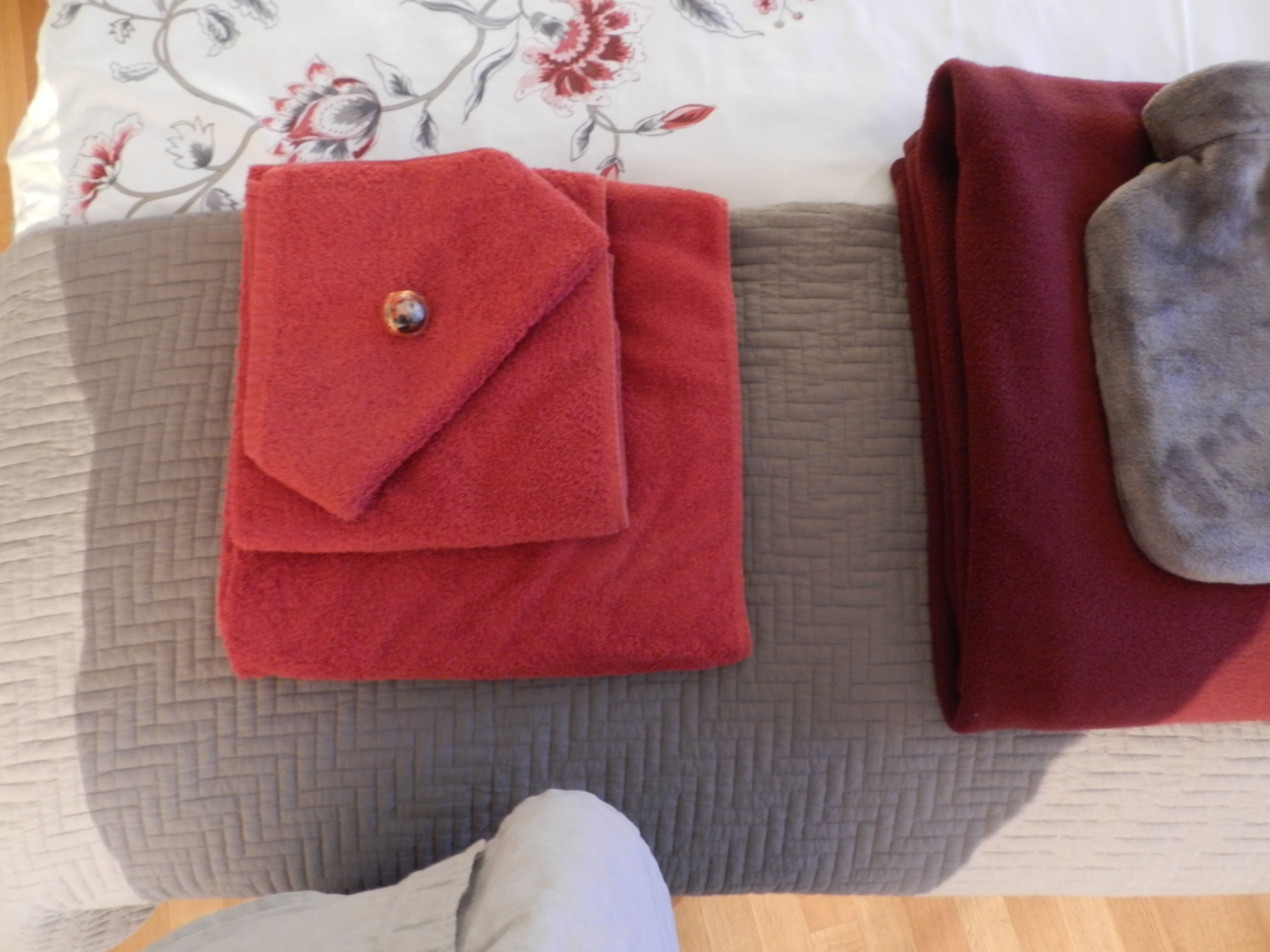 A neatly folded red towel with a decorative button on a bed with a floral-patterned pillow and a grey quilted blanket.