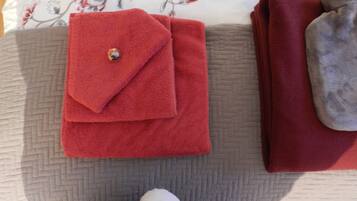A neatly folded red towel with a decorative button on a bed with a floral-patterned pillow and a grey quilted blanket.