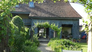 Une maison de campagne avec une entrée donnant sur le jardin, une véranda en bois et une petite table accompagnée d’une chaise.