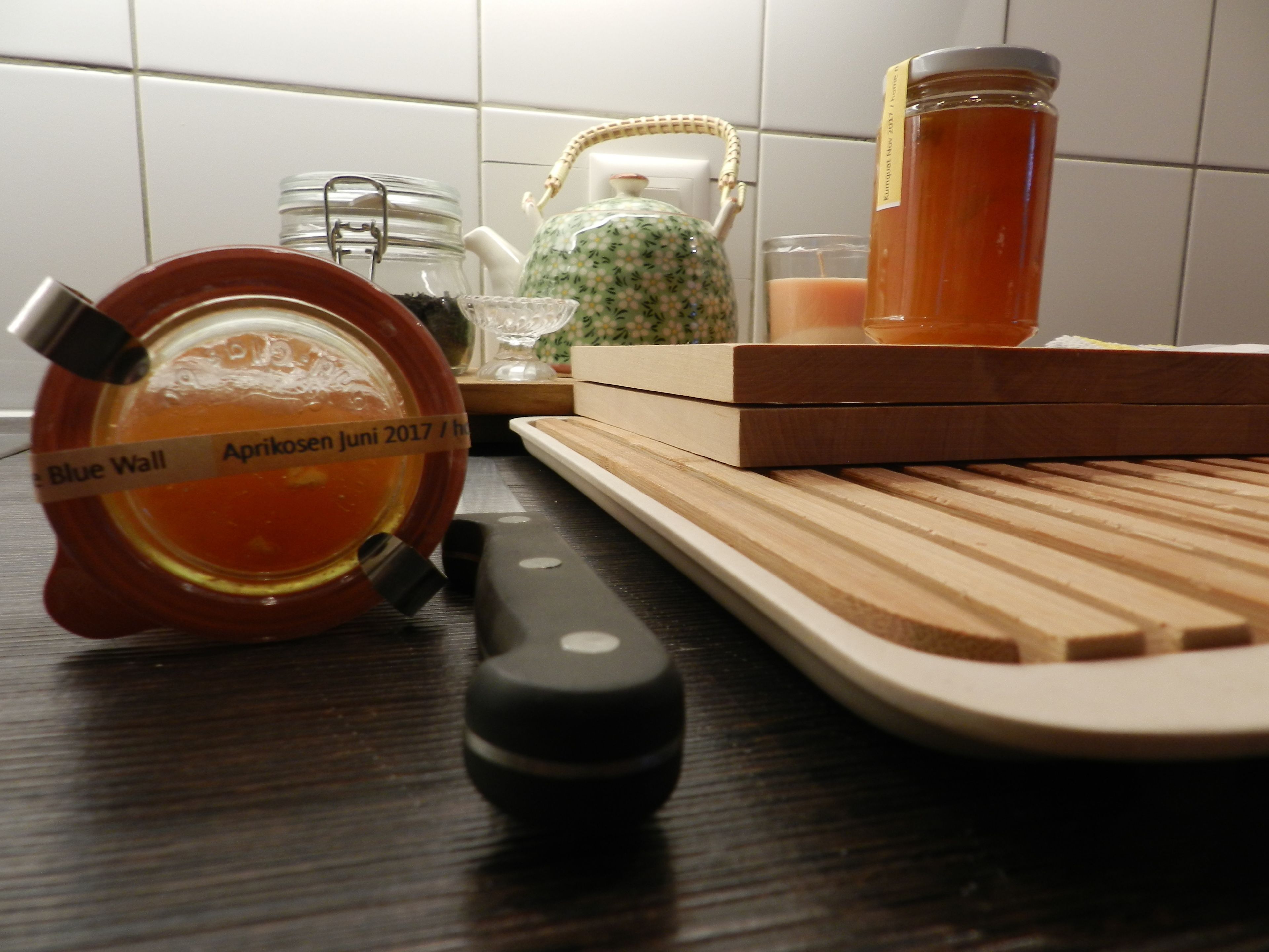 A kitchen counter with a wooden cutting board, a knife, a jar of honey, a teapot, and a glass jar.