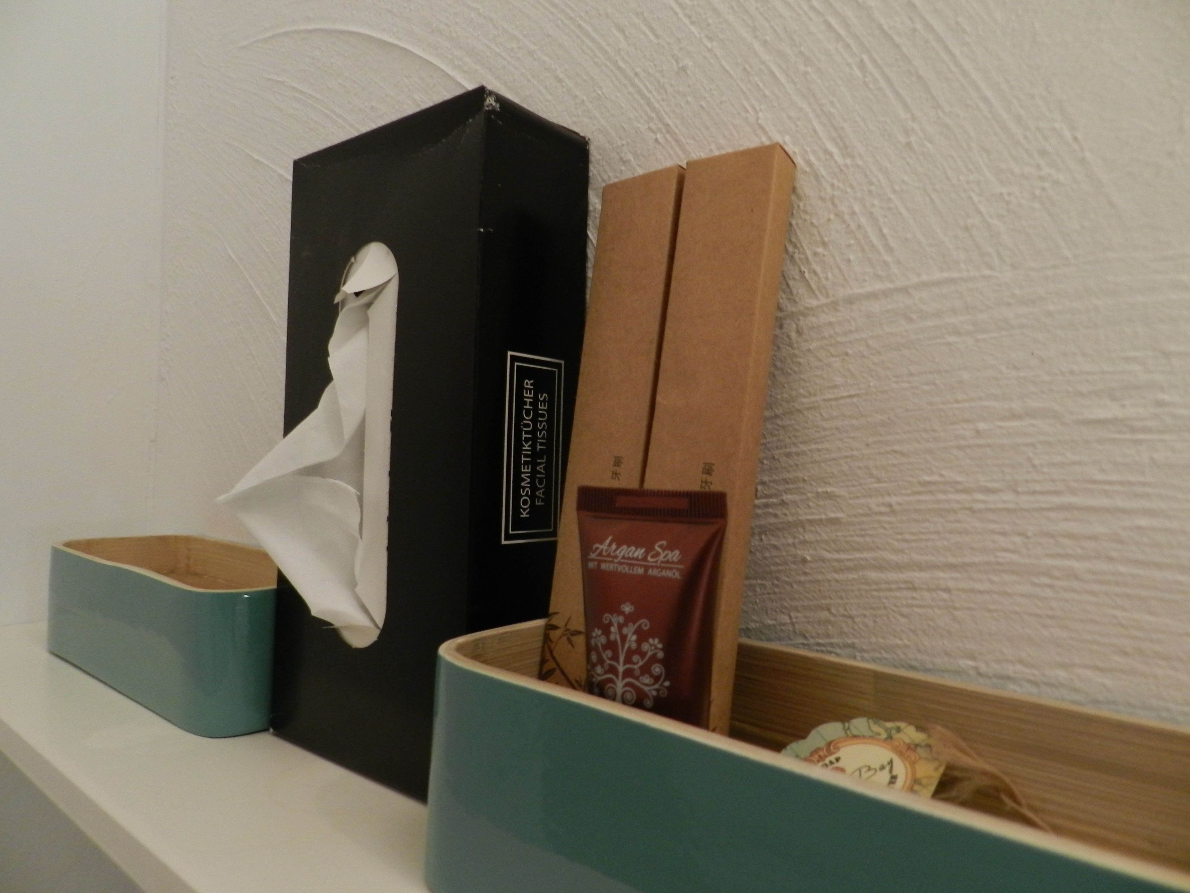 A shelf with a black box labeled 'KOSMETIKTÜCHER FACIAL TISSUES', a brown box labeled 'Argan Son', and a blue container with a white tissue.