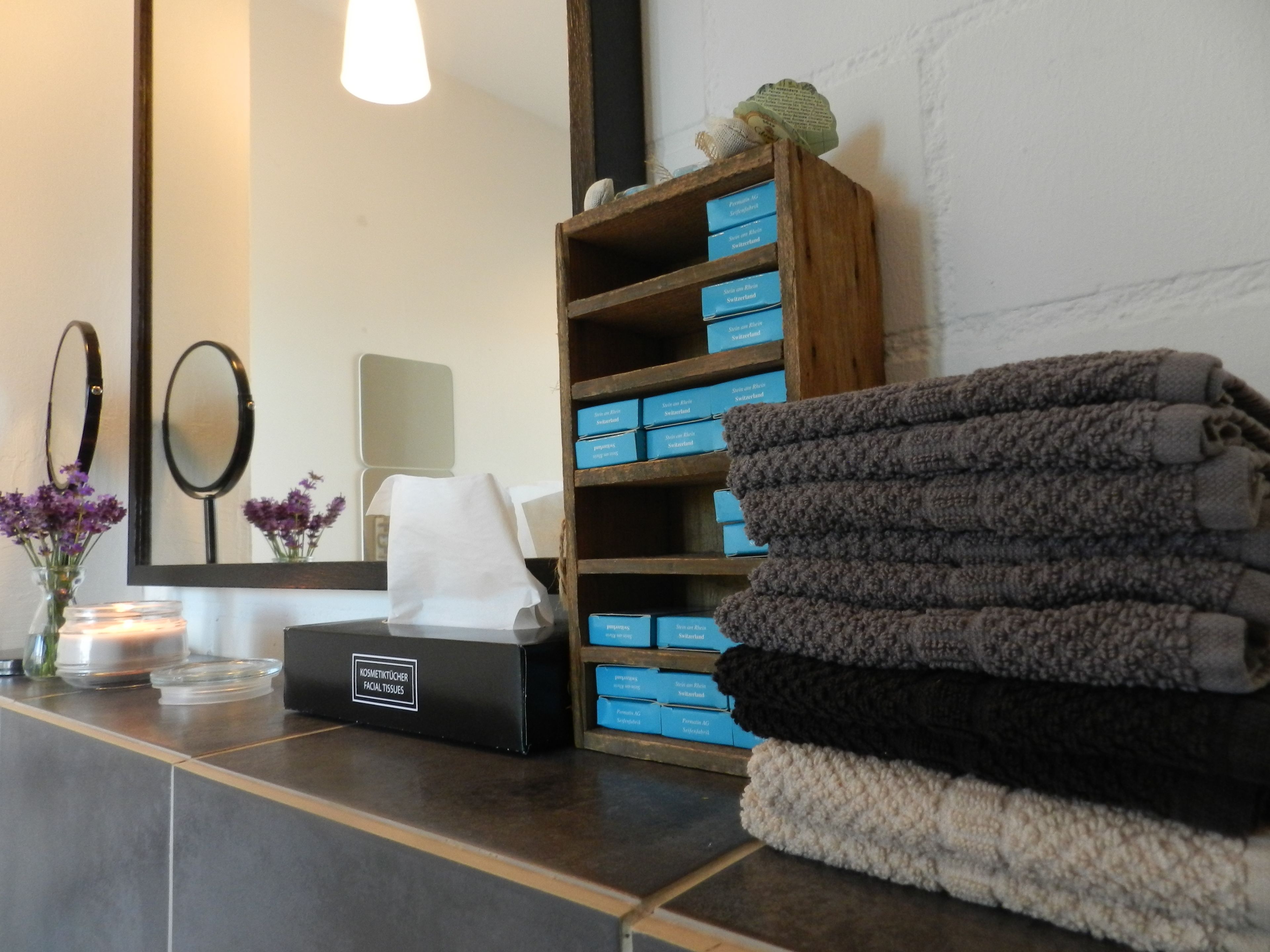 Een badkamer met een spiegel, een plank met blauwe dozen, een stapel handdoeken en een doos met de tekst 'KOSMETIKTÜCHER FACIAL TISSUES'.