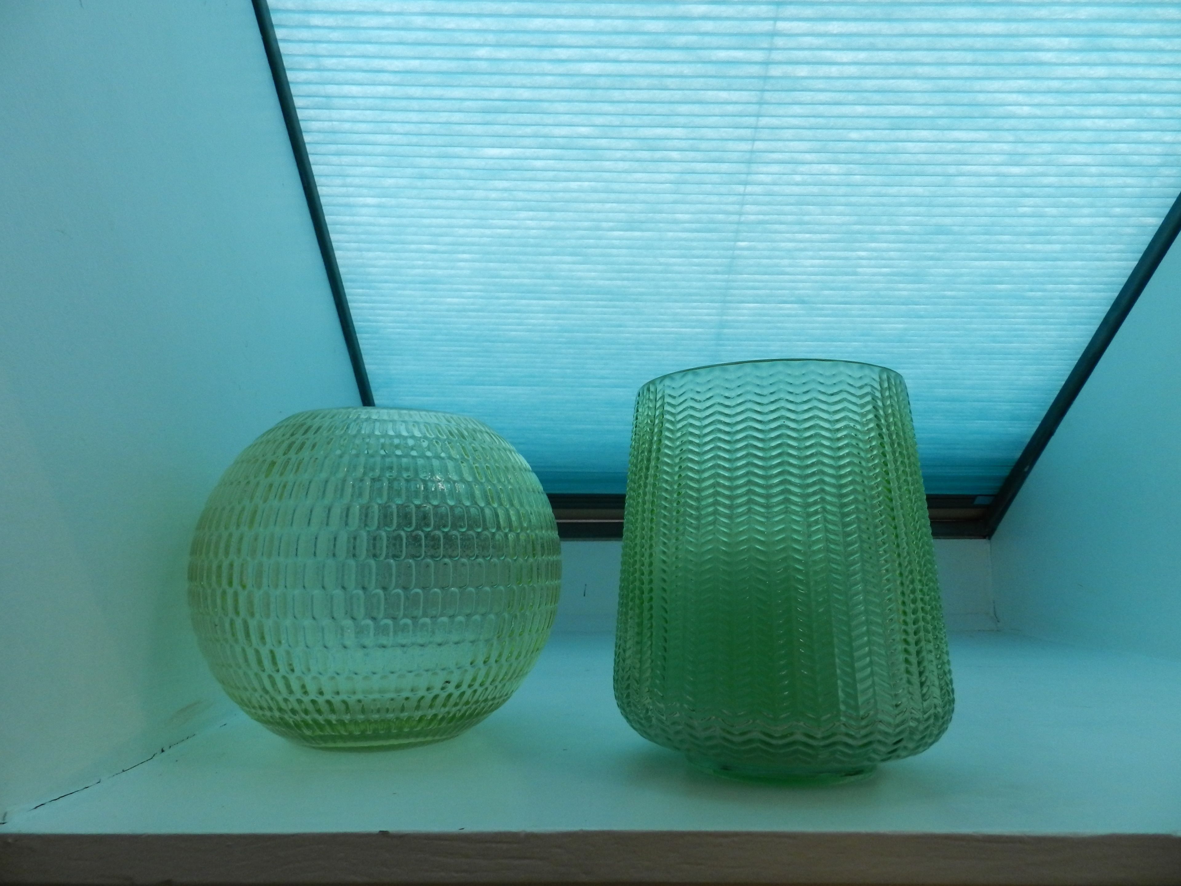 Two glass vases on a shelf under a window with blinds.