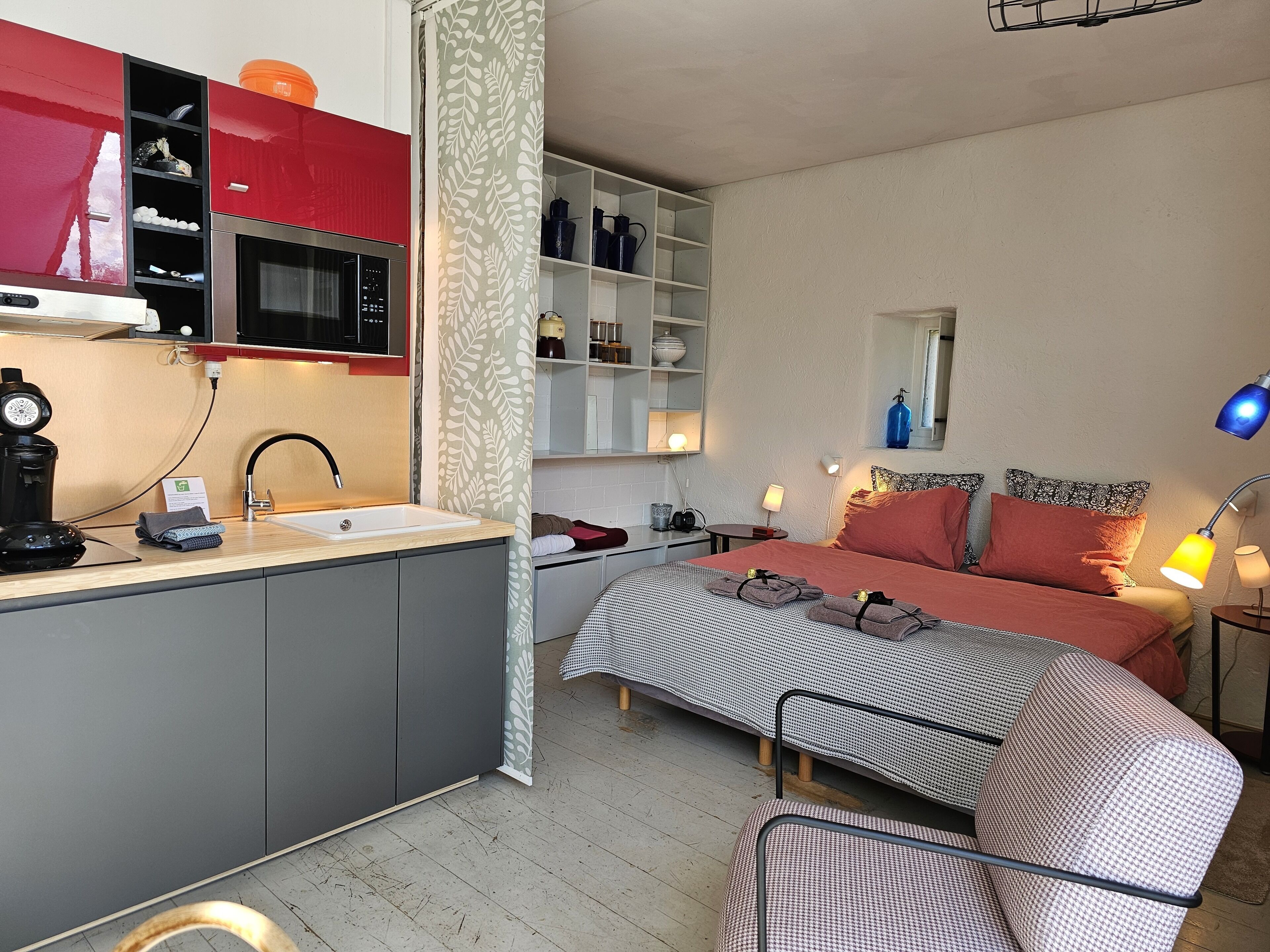 A modern hotel room with a kitchenette, a bed with patterned bedding, bedside lamps, and a small table with a lamp.
