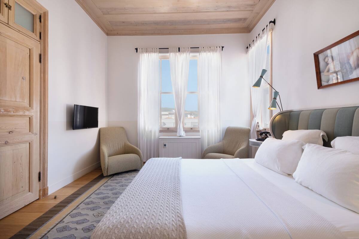 A hotel room with a bed, two armchairs, a TV, a window with curtains, a lamp, and a painting on the wall.