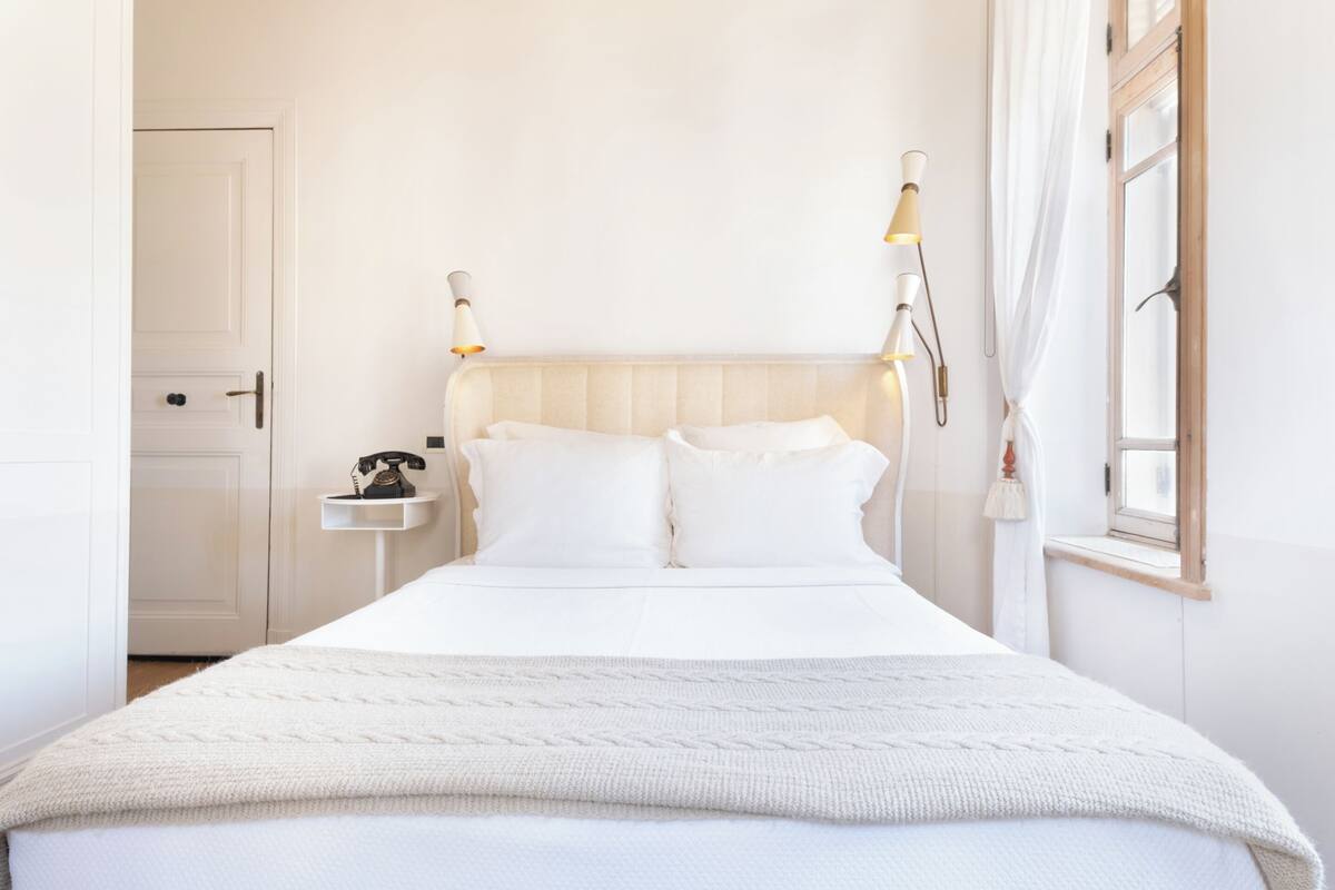 A neatly made bed with a white headboard, a bedside table with a lamp, and a window with curtains.