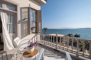 A balcony with white furniture, a table with a vase of flowers, and a view of the sea and distant mountains. - Nars Ilica Hotel Beach (Cesme)