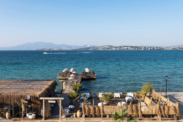 A coastal restaurant with outdoor seating overlooking the sea, a wooden deck, and a thatched-roof structure. - Nars Ilica Hotel Beach (Cesme)
