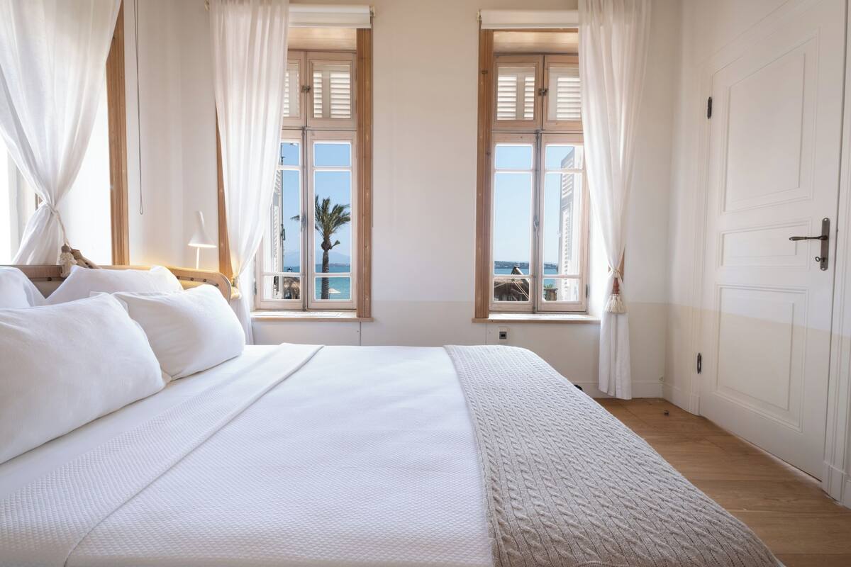 A bedroom with a large bed, two windows with shutters, a view of the sea, and a door.