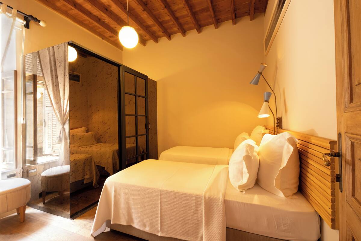 A bedroom with two beds, a wooden ceiling, a window with curtains, and a bedside lamp.