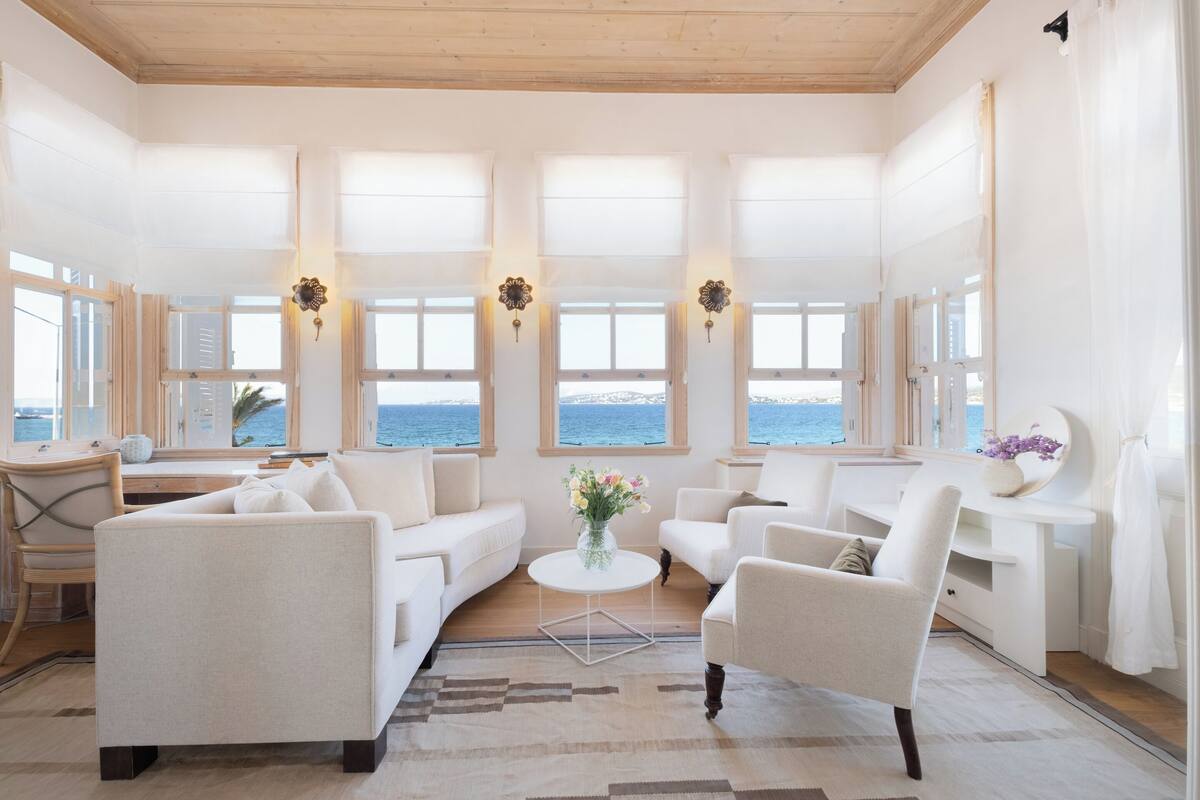 A spacious living room with a large window overlooking the sea, a white sofa, a coffee table, and a desk with a lamp.