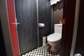 A small, compact bathroom with a toilet, a hand dryer, and a trash can.
