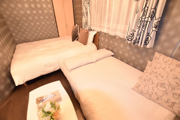 A hotel room with two single beds, a wooden headboard, a small table with a floral table runner, and patterned curtains. - Mell-Theater 1102 (Osaka)