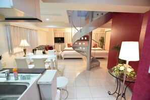 A modern hotel room with a kitchen area, a dining table, a sofa, a TV, and a spiral staircase.