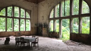 An abandoned room with a table and chairs, large arched windows, and a view of greenery outside.