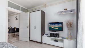 Una sala moderna con televisor, armarios blancos, una zona de comedor con sillas y una cama.